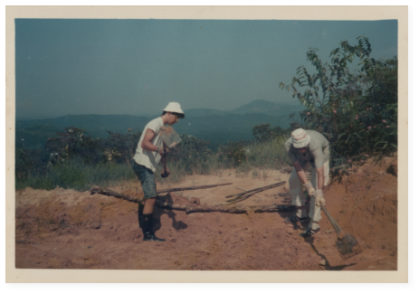 Two men with cangkuls (local shovels) preparing the ground for planting before a mountainous background.