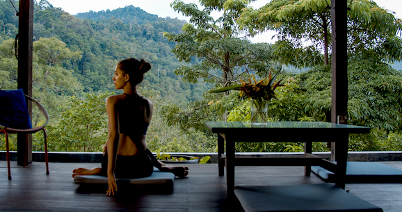 A woman is sitting on a yoga mat in a room with a view of houses in the forest.